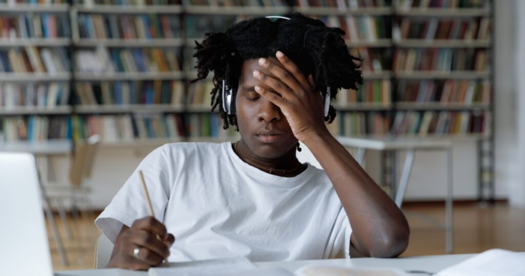 African student feels tired due to long preparations for college exam, deadline task, suffers from headache sit at table with laptop in library. Information overload during admission, stress concept