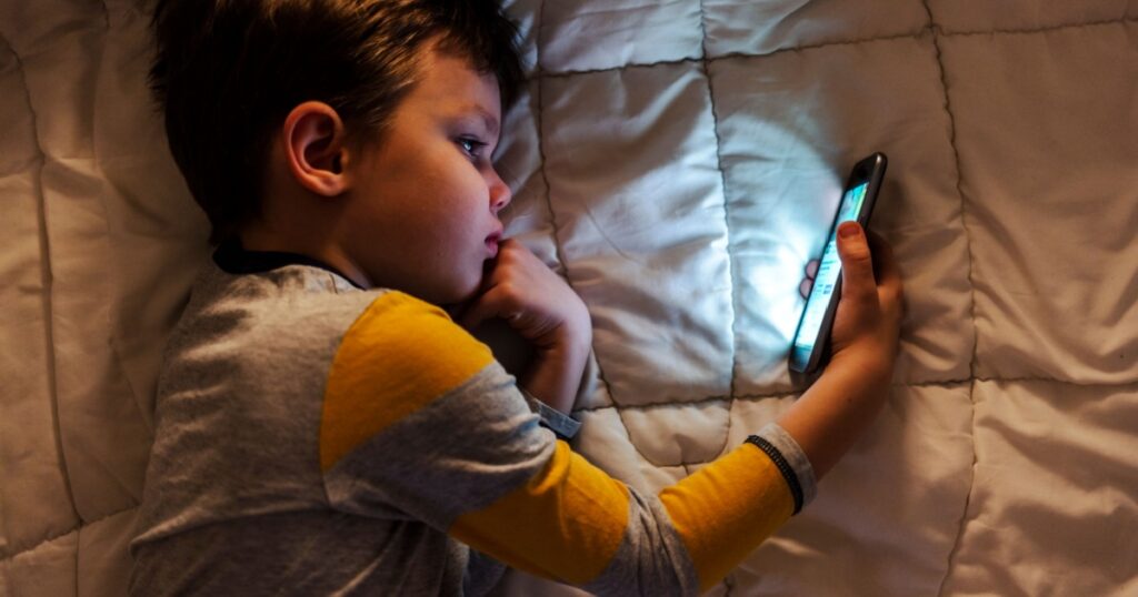 Cute little boy using a smartphone. Kid playing with mobile phone, lying on a bed. Freetime. Technology and internet concept. Smiling toddler laying on bed and looking at smartphone. Light reflectio