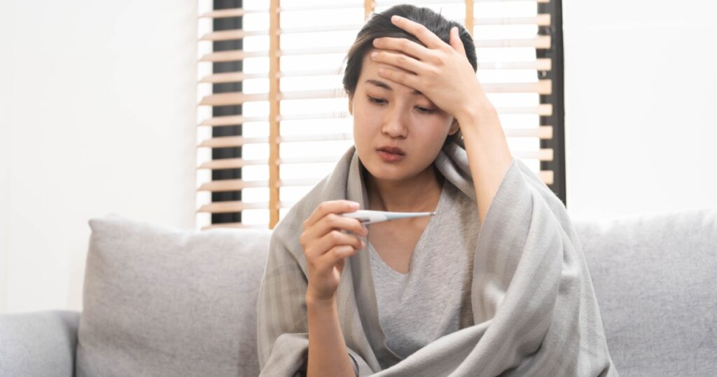 Sick, influenza asian young woman, girl headache have a fever, flu and check thermometer measure body temperature, feel illness sitting on sofa bed at home. Health care person on virus, covid-19.