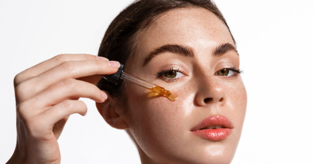 Beauty woman with freckles, naked shoulders and nude face, apply serum vitamin c, argan oil tea tree ingredients in pippette dropper under eye, treating skin for nourishing and moisturizing effect