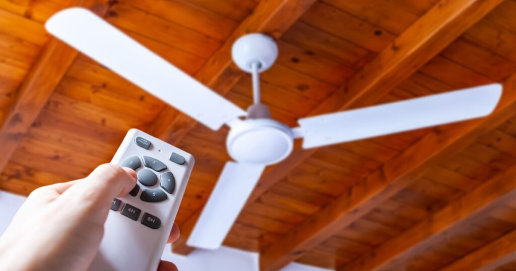 A man uses a remote control to turn on a white ceiling fan mounted in a house with wooden ceilings.