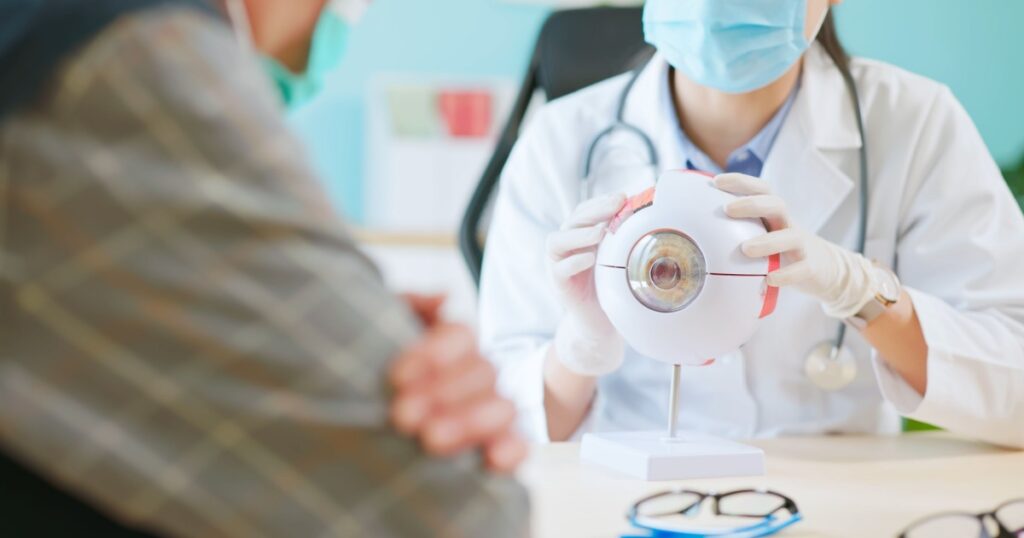 close up of asian female doctor wearing face mask is showing eyeball model and explaining about eyes disease to elder senior man patient in hospital