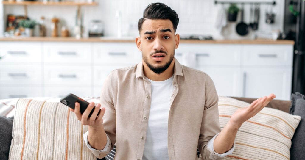 Confused puzzled indian or arabian guy in casual clothes, sits on a sofa in an interior living room, holds a smartphone in his hand, looks questioningly at the camera, spreading his arms around