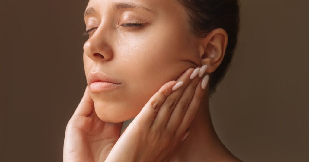 Cropped shot of young caucasian woman touching skin with hands massaging her face isolated on a dark brown background. Rejuvenation, facelift, facefitness. Cosmetology and beauty concept