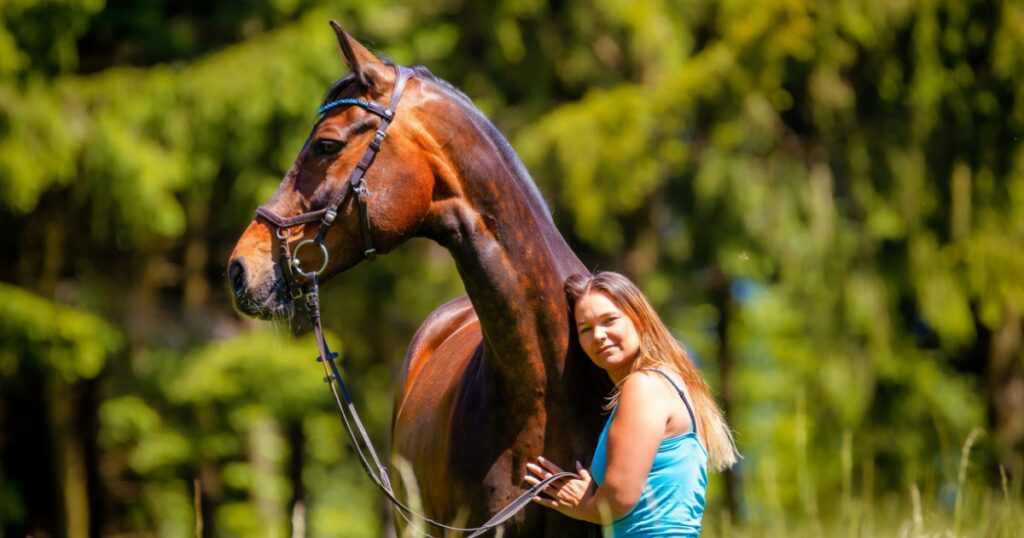 Horse stands with a young woman in a meadow, horse looks attentively to the right and the woman snuggles up to the horse's neck.