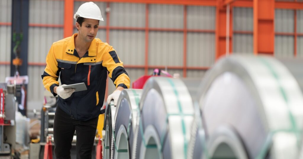 Metalwork manufacturing, warehouse of raw materials. Male factory worker inspecting quality rolls of metal sheet in factory during manufacturing process, wearing safety uniform, use digital tablet