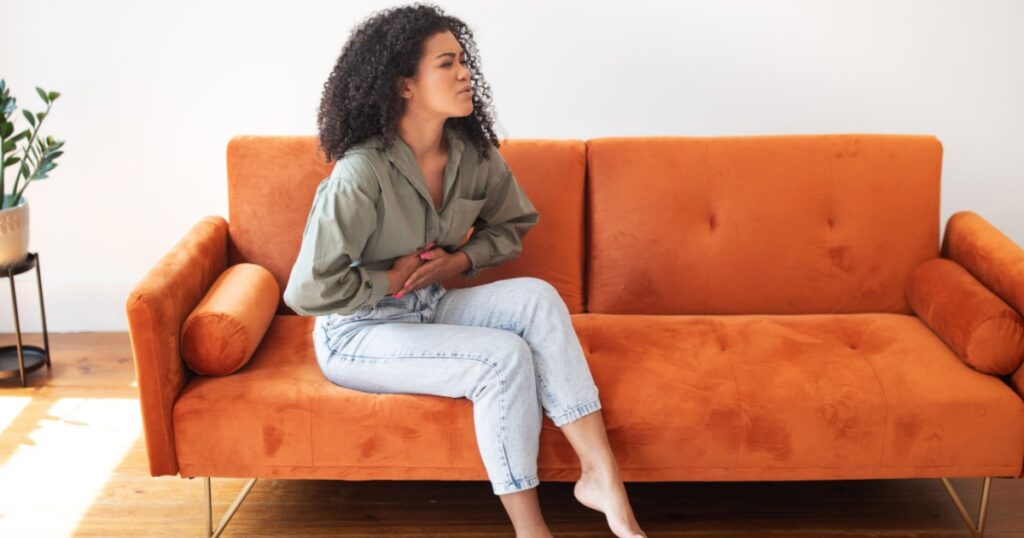 Gastrointestinal Distress. Young brazilian woman at home dealing with abdominal discomfort, touching aching belly, showcasing challenges of digestive issues and period pain, sitting on couch indoor