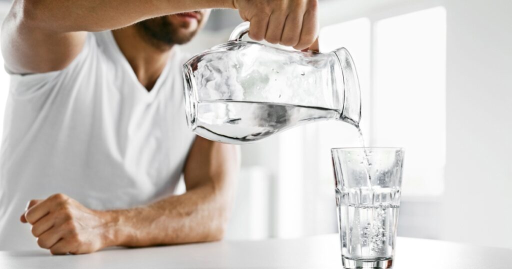 Drink Water. Close Up Of Handsome Young Man Pouring Fresh Pure Water From Pitcher Into A Glass In Morning In Kitchen. Beautiful Athletic Male Model Feeling Thirsty. Healthy Nutrition And Hydration