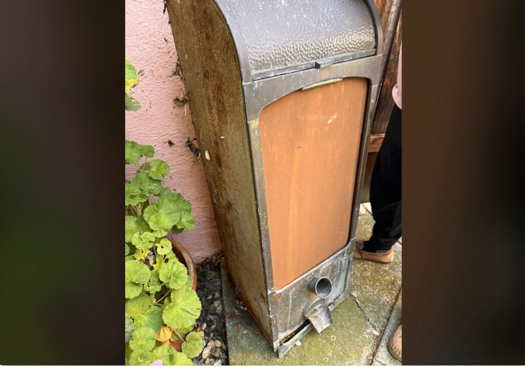 Wooden box with metal casing and spout. Feet and a plant in the background.