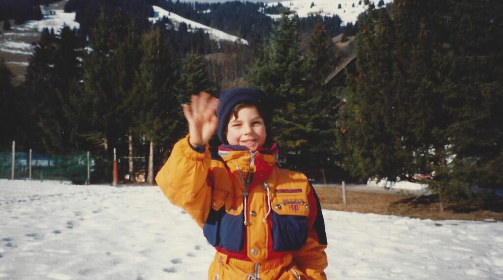 Young Carlo Acutis waving to the camera, bundled up in winter gear. Snow and trees in the background.
