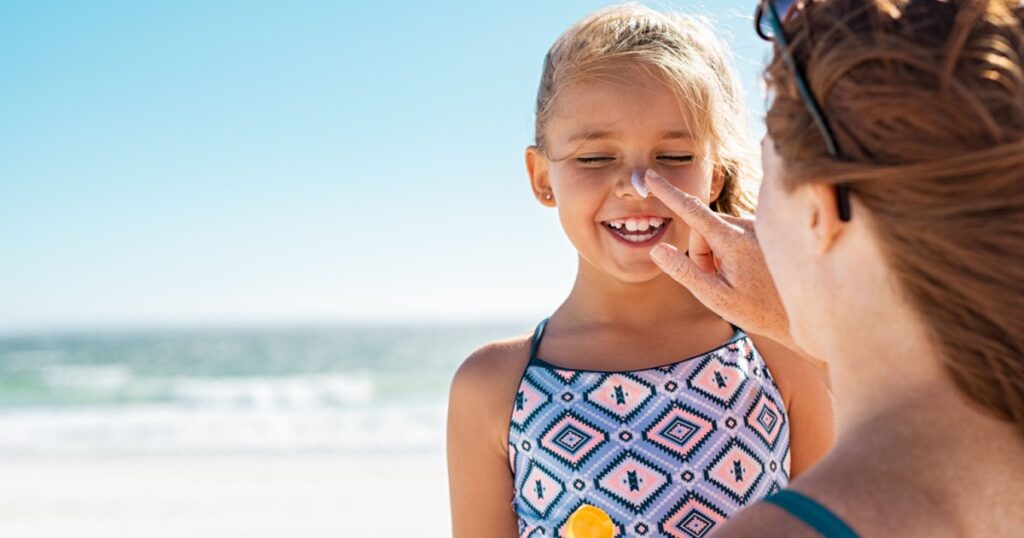 Young mother applying protective sunscreen on daughter nose at beach. Woman hand putting sun lotion on child face. Cute little girl with sunblock at seaside with copy space.
