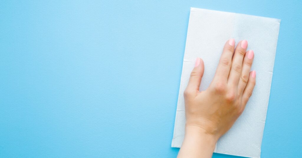 Woman's hand wiping pastel blue desk with white paper napkin. General or regular cleanup. Close up. Empty place for text or logo.