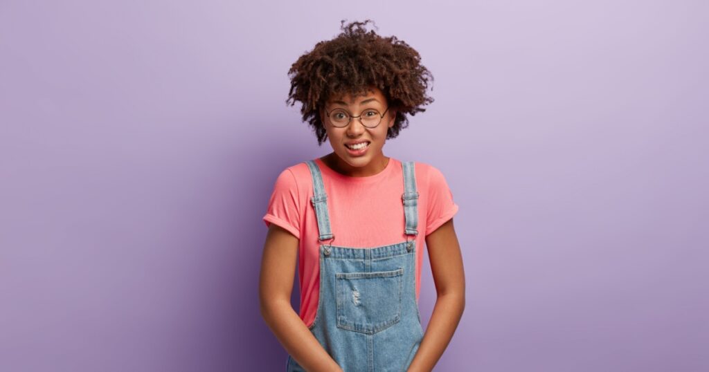 Unhappy dark skinned woman holds crotch, needs toilet, has problematic situation, wears pink t shirt and denim sarafan, suffers from cystitis, isolated over purple background. People and urgency
