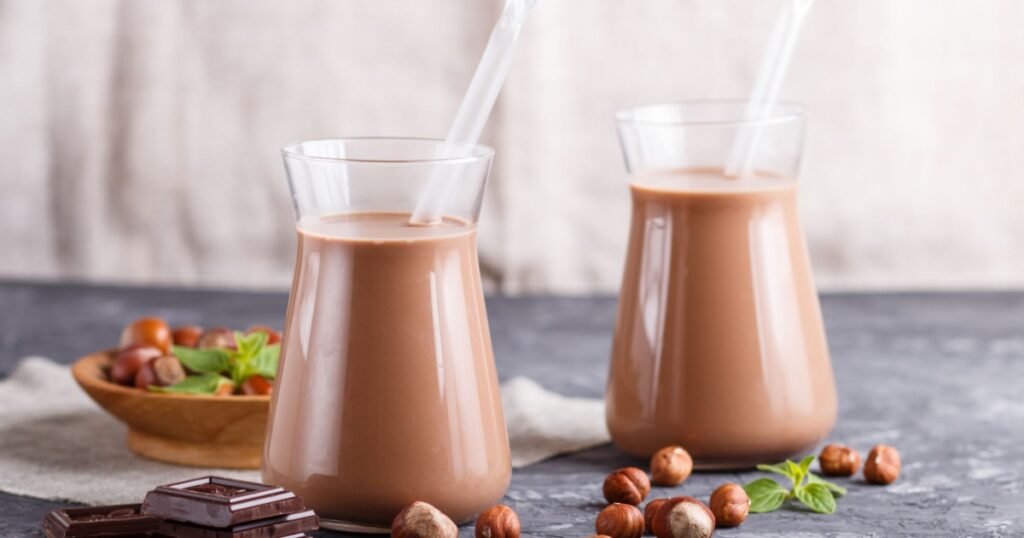 Organic non dairy hazelnut chocolate milk in glass and wooden plate with hazelnuts on a black concrete background. Vegan healthy food concept, close up, side view.
