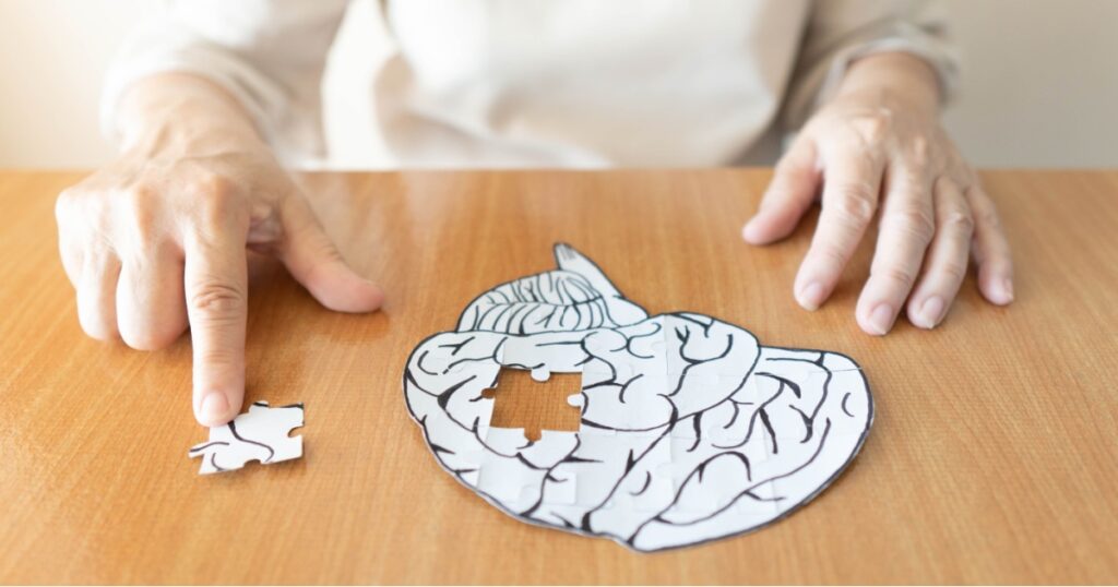 Elderly woman hands putting missing white jigsaw puzzle piece down into the place as a human brain shape. Creative idea for memory loss, dementia, Alzheimer's disease and mental health concept.