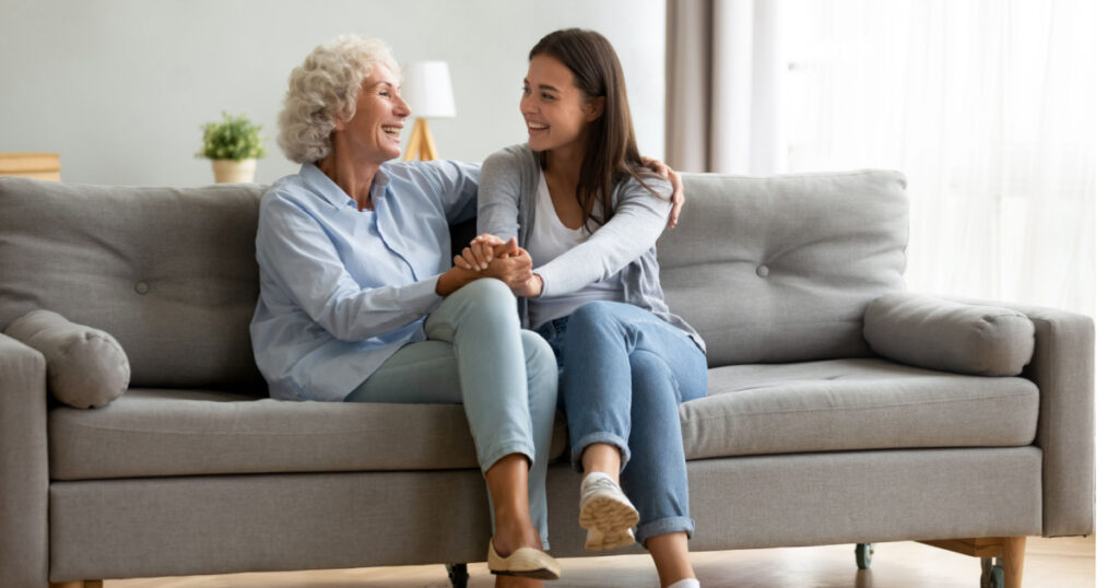 Happy adult granddaughter and senior grandmother having fun enjoying talk sit on sofa in modern living room, smiling old mother hugging young grown daughter bonding chatting relaxing at home together