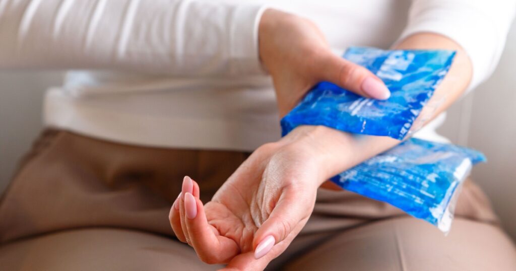 Close up of woman applying cold compress to a her painful wrist caused by prolonged work on the computer, laptop, selective focus on hand. Carpal tunnel syndrome, arthritis, neurological disease.