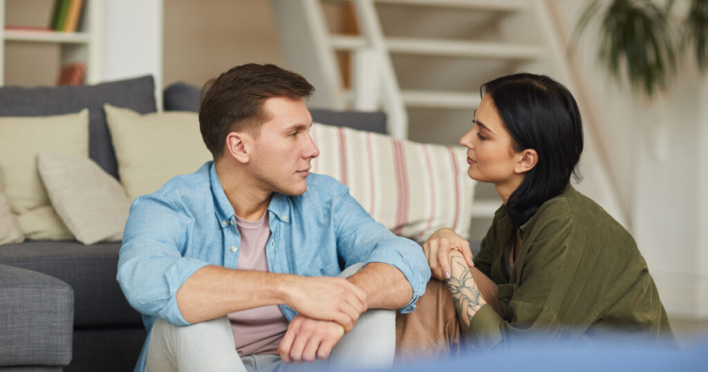 Warm toned portrait of modern young couple talking to each other sincerely while sitting on floor in cozy home interior, copy space