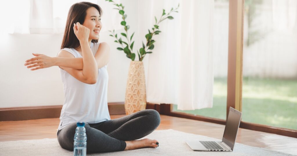 Smiling Asian woman doing yoga shoulder stretching online class from laptop at home in living room. Self isolation and workout at home during COVID-19.