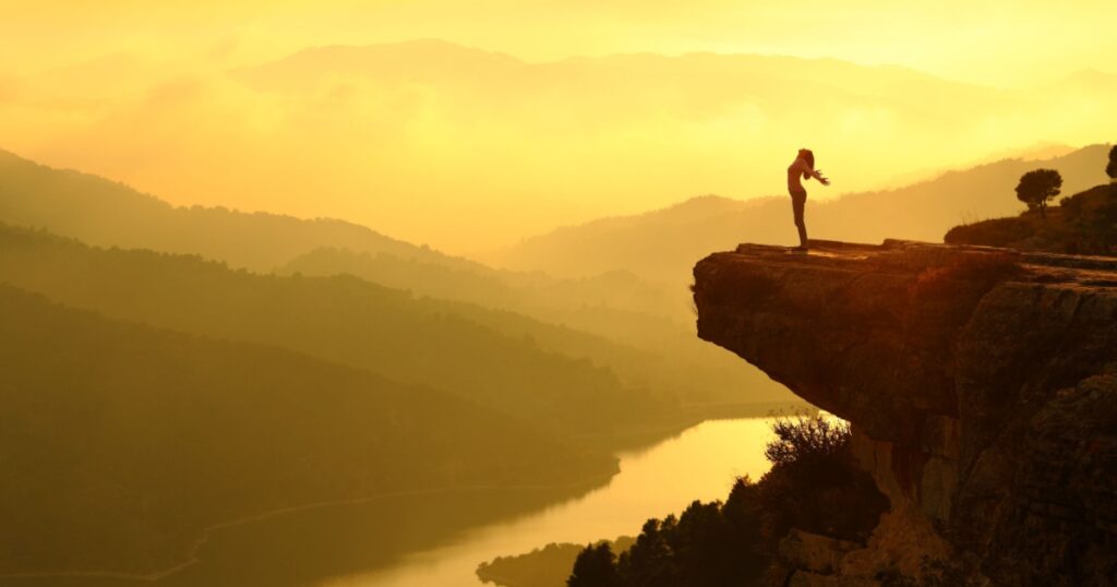 Woman silhouette breathing fresh air in the top of a cliff in the mountain at sunset with a beautiful landscape in the background