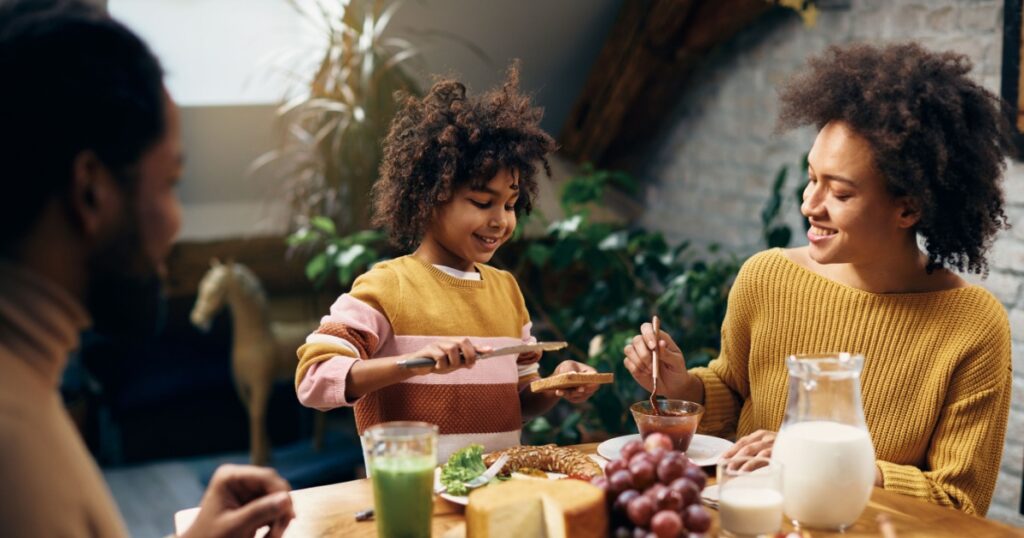 Happy black girl spreading butter on toasted bred while eating breakfast with her parents in dining room.