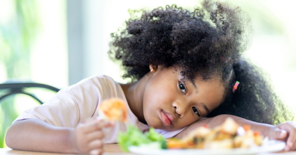 A young, curly haired African American girl sits bored looking at food. Do not want to eat food. Childhood concepts and healthy eating.