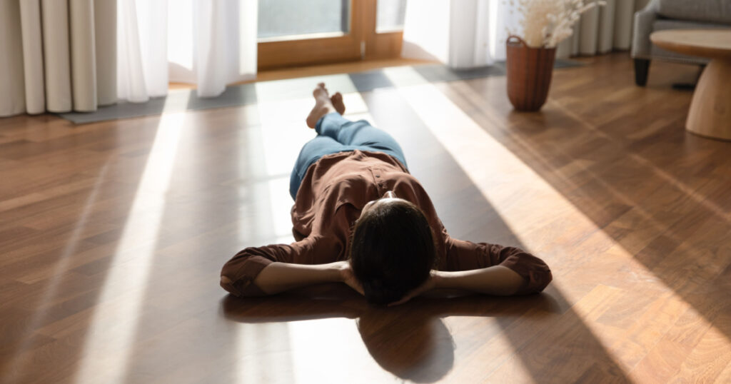 Relaxed happy young indian ethnicity woman homeowner lying alone on warm wooden floor with underfloor heating, enjoying carefree peaceful weekend leisure time alone in modern stylish living room.