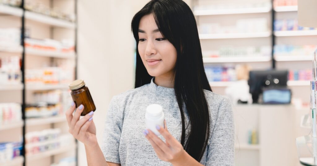 Female asian customer client looking for medications, medicines, comparing two jars with drugs, pills, vitamins, antibiotics, painkillers, choosing between two products in pharmacy drugstore