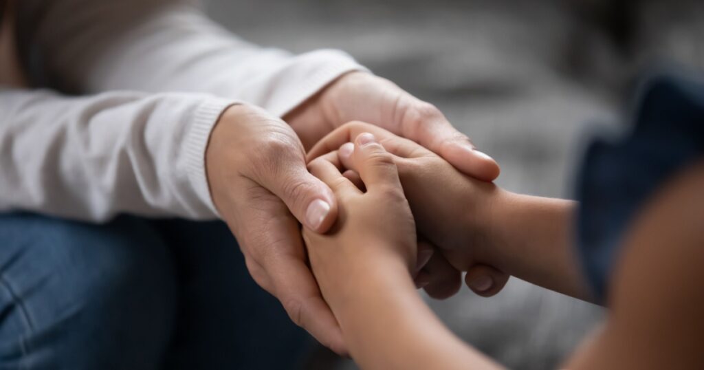 Close up compassionate young foster parent holding hands of little kid girl, giving psychological help, supporting at home. Sincere different generations family sharing secrets or making peace.
