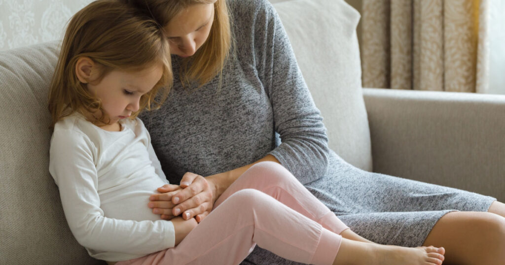 Child sitting on sofa and suffering from stomach ache at home room. Young adult mother hand touching daughter painful belly and trying calm down.