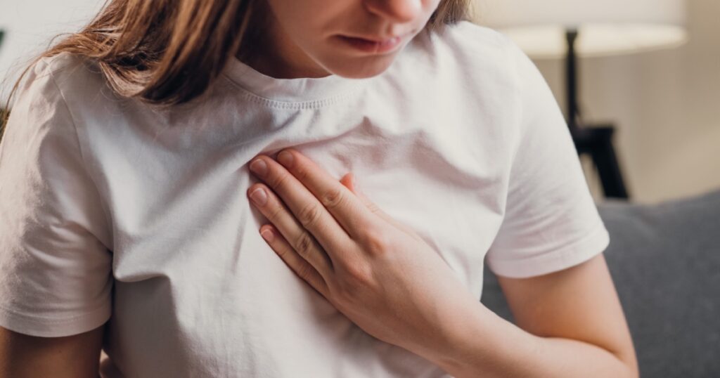 Young unhealthy female sit on couch having difficulty breathing pain of heart, touches his chest with hand. Trouble breathing, chest pain. Heart attack, thoracic osteochondrosis, panic attack concept