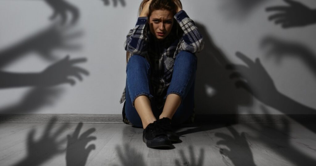 Paranoid delusion. Scared woman sitting near wall. Shadows of hands reaching for her symbolizing fear and anxiety