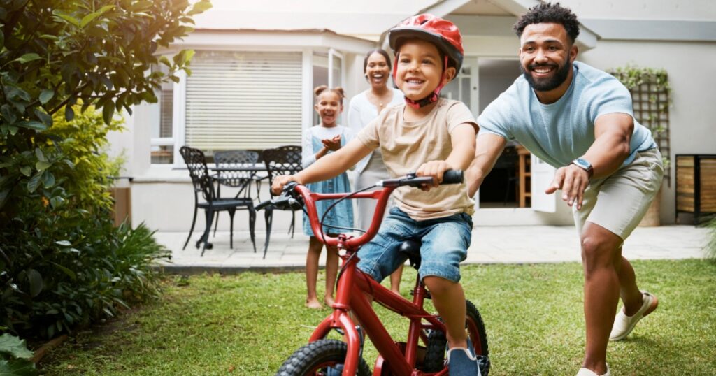 Learning, bicycle and proud dad teaching his young son to ride while wearing a helmet for safety in their family home garden. Active father helping and supporting his child while cycling outside