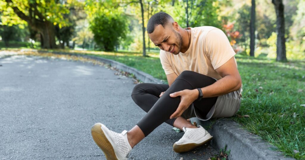 Runner injured his leg while jogging in the park near the trees, hispanic man has muscle spasm massaging his knee, man is running and exercising