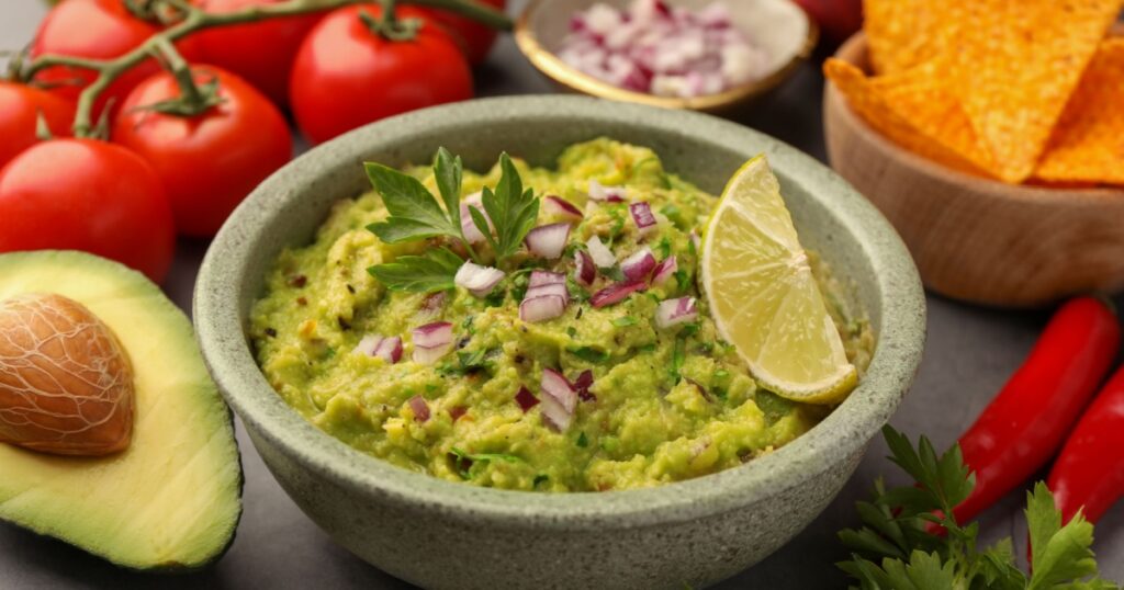 Bowl of delicious guacamole, nachos chips and ingredients on grey table, closeup
