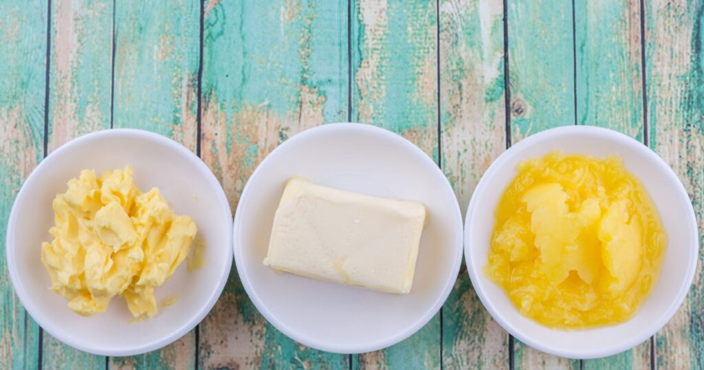 A block of butter, margarine and ghee in white bowls over rustic wooden background
