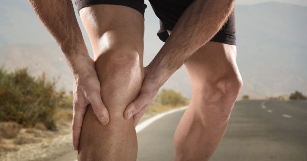 young sport man with strong athletic legs holding knee with his hands in pain after suffering muscle injury during a running workout training in asphalt road in muscular or ligament wound