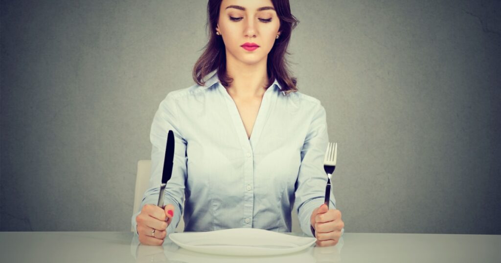 Serious woman with fork and knife sitting at table with empty plate isolated on gray wall background