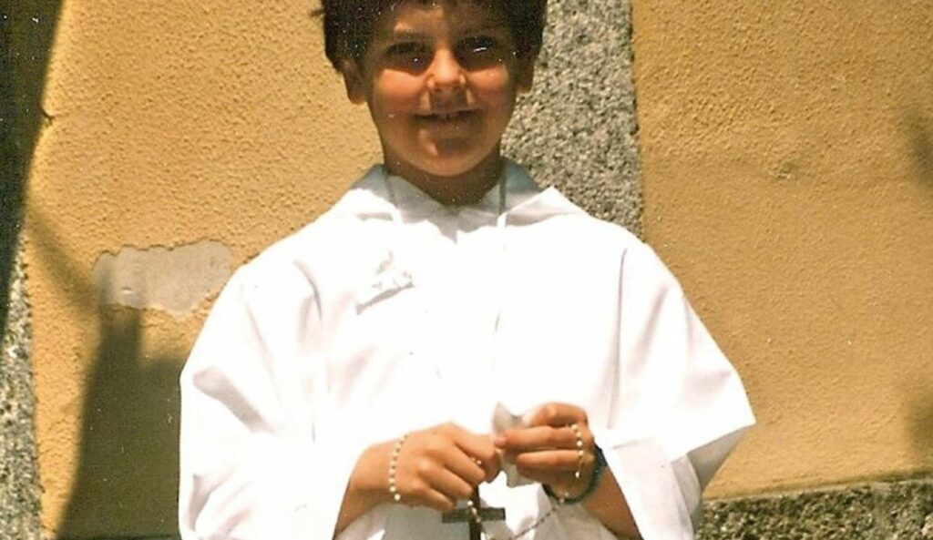 Carlo Acutis wearing traditional Catholic Church attire and holding a rosary. Building in the background.