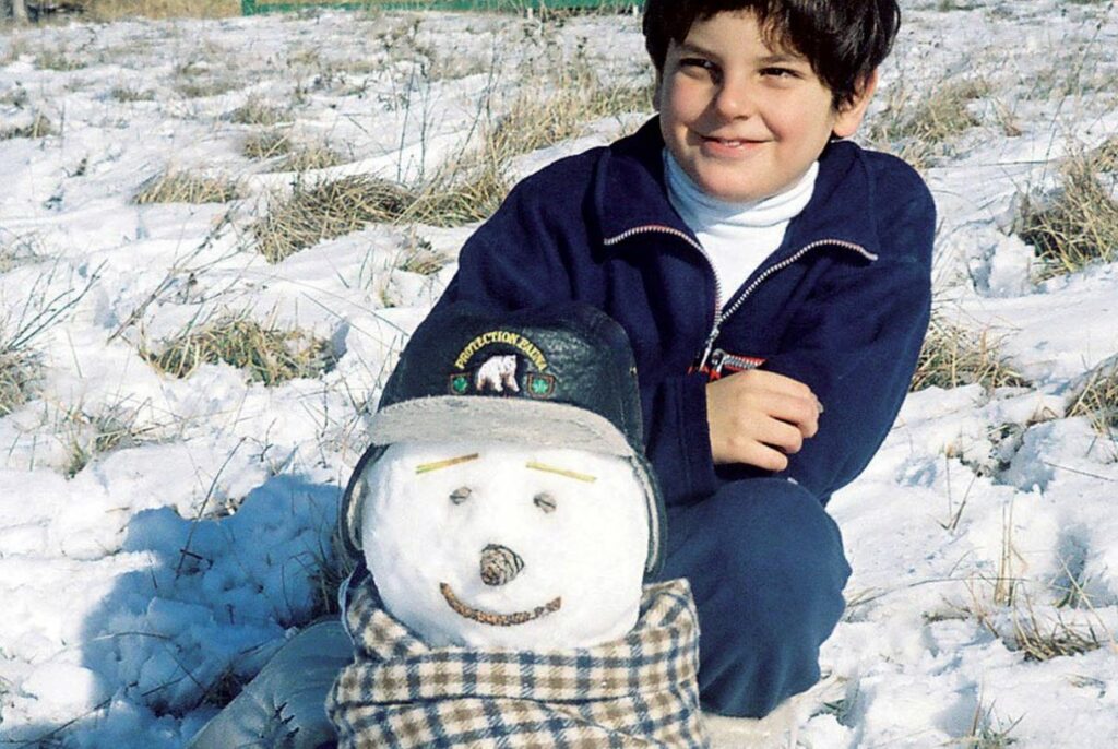 Carlo Acutis poses with a snowman wearing a hat and scarf. Snow and grass in the background.