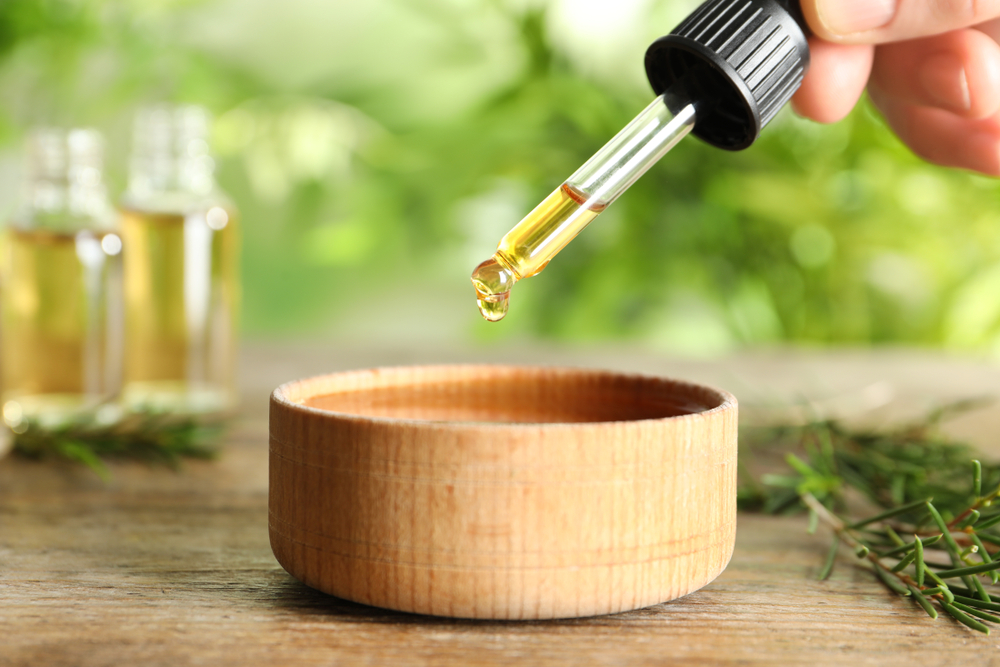 Woman dripping natural tea tree oil in bowl on table, closeup
