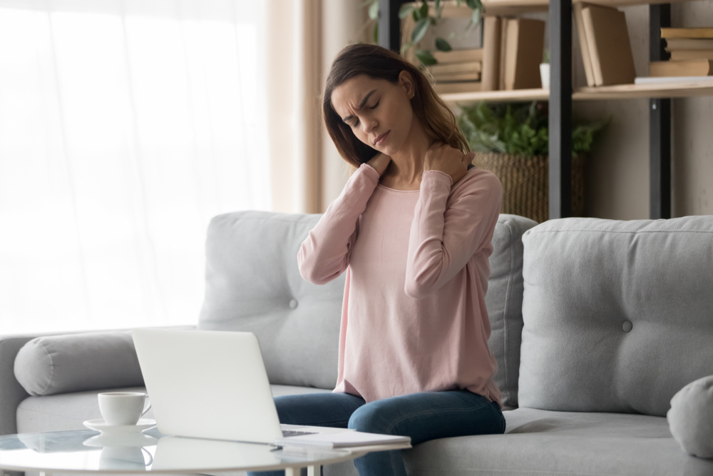 Tired young woman touch stiff neck feeling hurt joint back pain rubbing massaging tensed muscles suffer from fibromyalgia ache after long computer work study in incorrect posture sit on sofa at home
