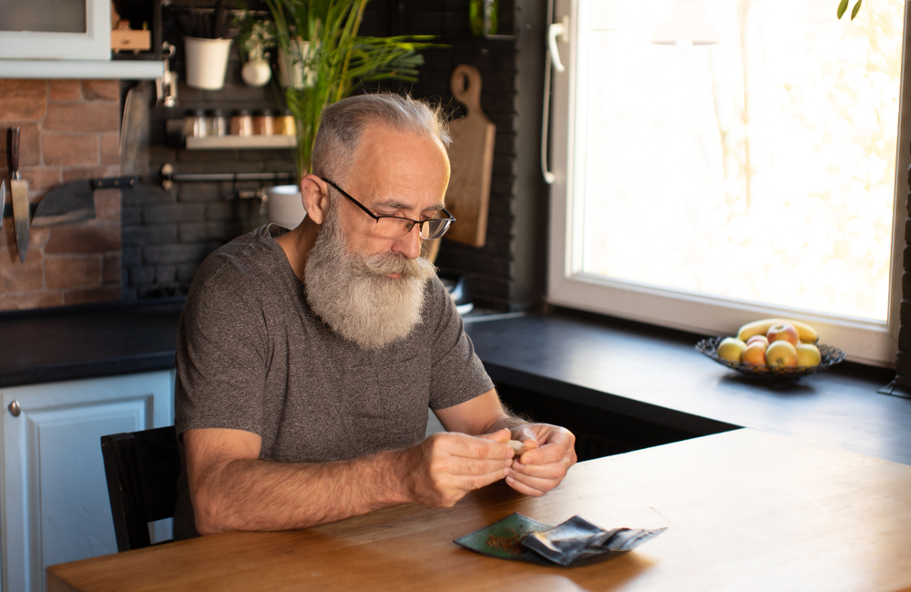 Bearded senior man preparing and rolling marijuana cannabis joint. Drugs narcotic concept.