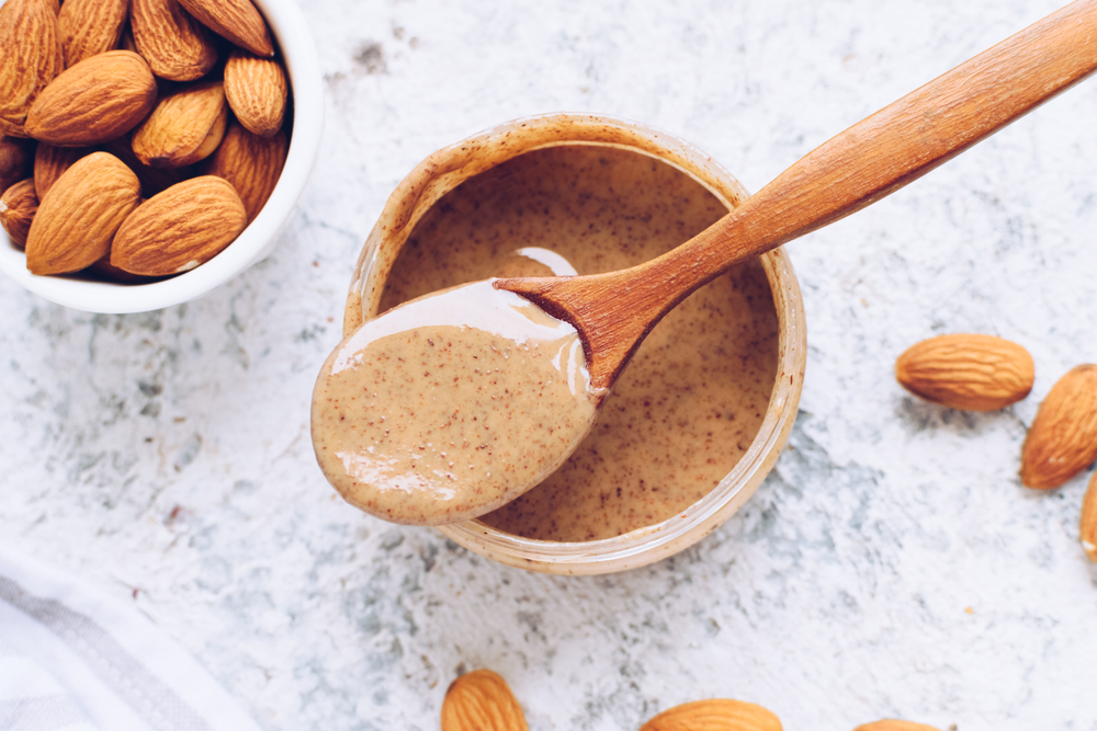 Almond nut butter in glass jar. Homemade raw organic almond nuts paste on grey background. Healthy natural food concept. Top view.
