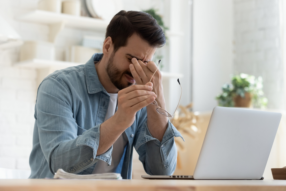 Tired young man feel pain eyestrain holding glasses rubbing dry irritated eyes fatigued from computer work, stressed man suffer from headache bad vision sight problem sit at home table using laptop
