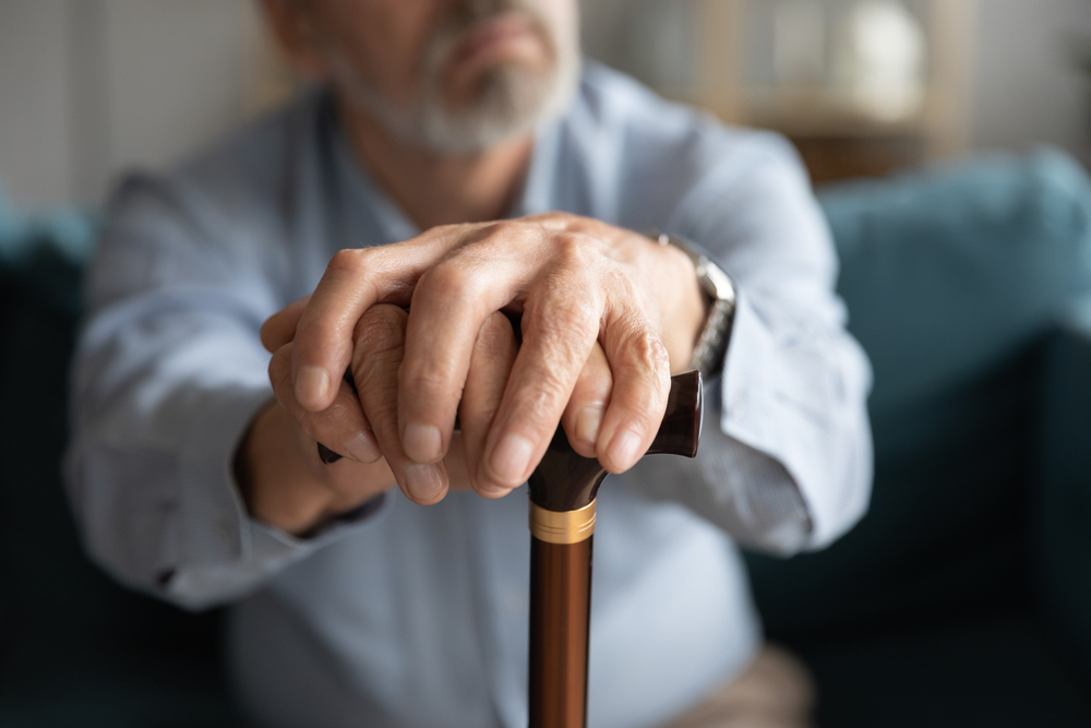 Close up of old man hands hold wooden cane look in distance thinking pondering, disabled mature 60s male feel lonely abandoned lost in thoughts missing mourning, elderly solitude concept