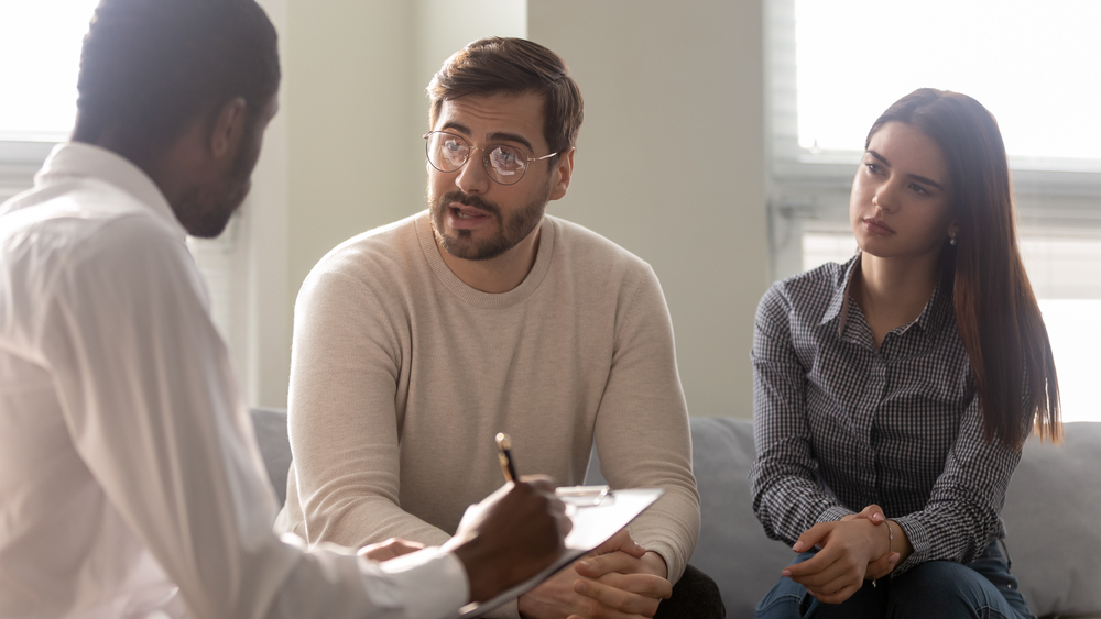 Disappointed couple at reception with family therapist. African american doctor listening man in bad mood. Family relationship disorder concept. Psychology session with coach
