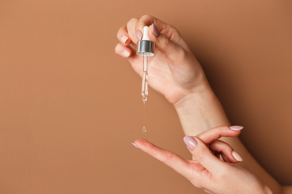 Hands of cropped white woman holding cosmetic serum pipette on the orange background.