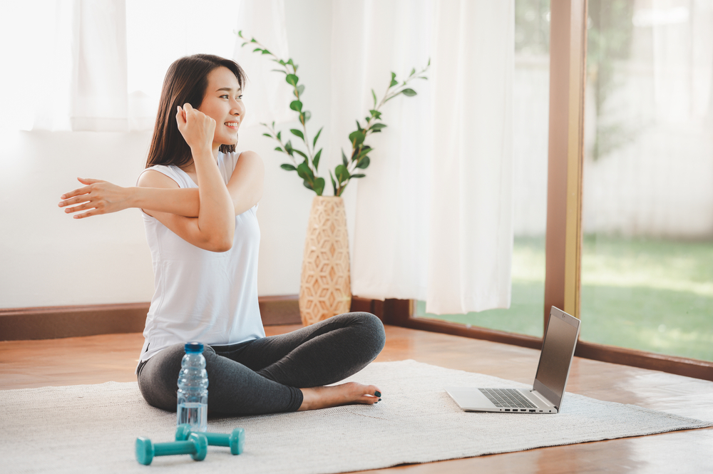 Smiling Asian woman doing yoga shoulder stretching online class from laptop at home in living room. Self isolation and workout at home during COVID-19.