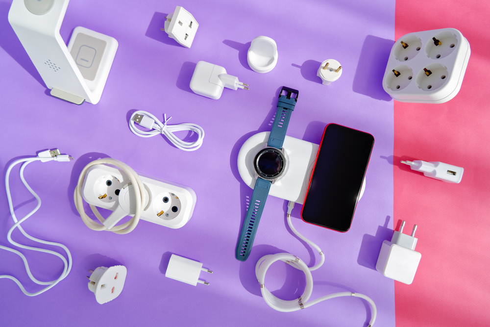 The concept of wireless charging of mobile gadgets. The smartphone and smartwatch are on the wireless charging pad. There are various adapters, cables and chargers nearby. Colored paper background.